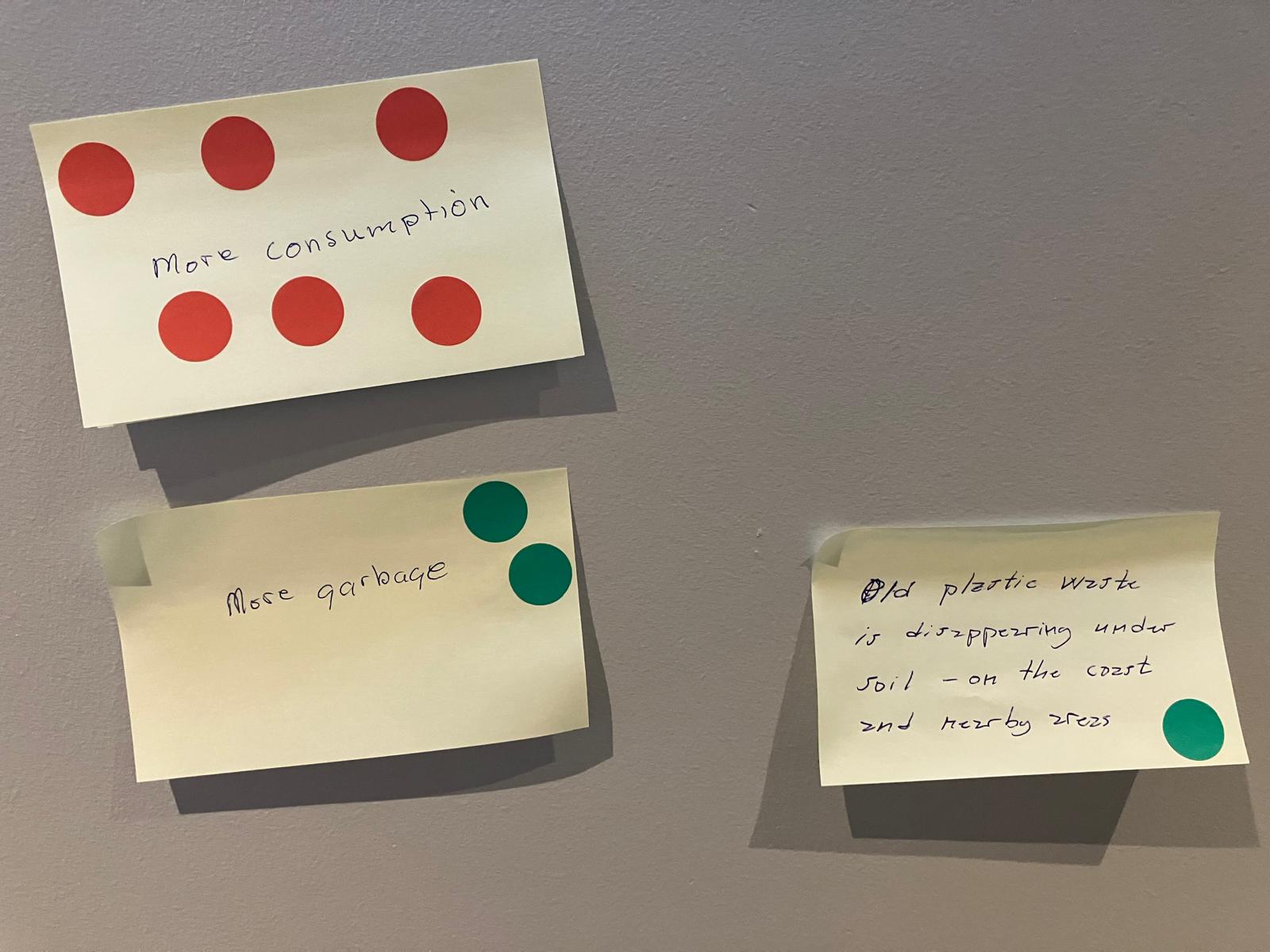 Post it notes on a wall, with text "more consumption", "more garbage" and "old plastic waste is disappearing under soil – on the coast and nearby areas". There are six red dot stickers on the first note that says "more consumption". There is one or two green dots on the latter two notes.