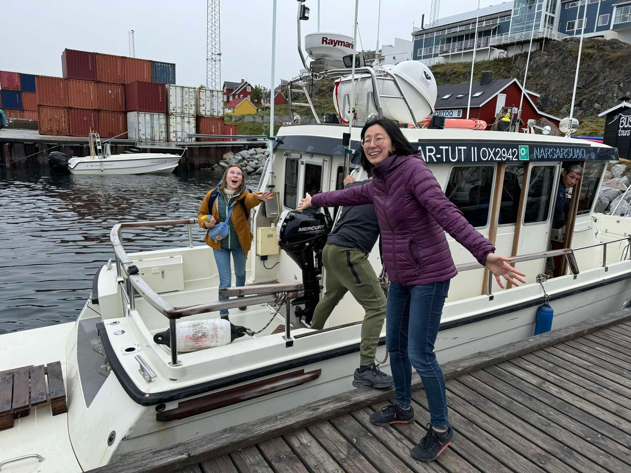 Tahnee Prior stands on a boat in a harbor and reaches humorously out to Christine Liang who stands on the pier and reaches out to Tahnee.