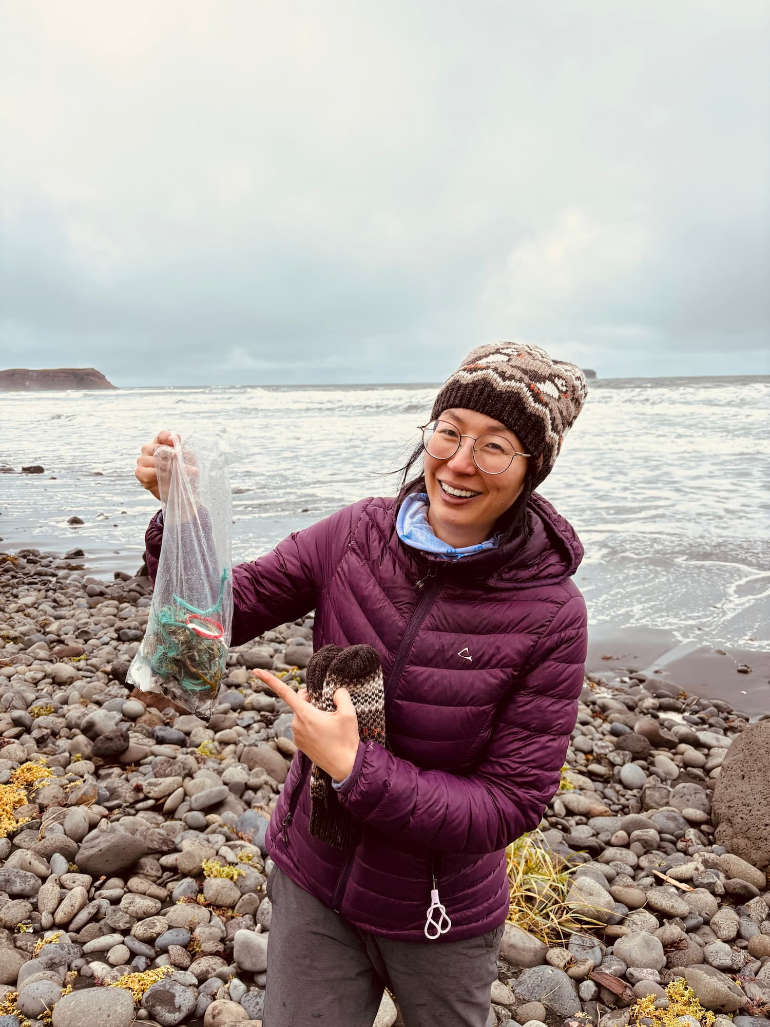 Christine Liang on a rocky beach in Iceland smiling for the camera and holding a little bag of plastic litter.