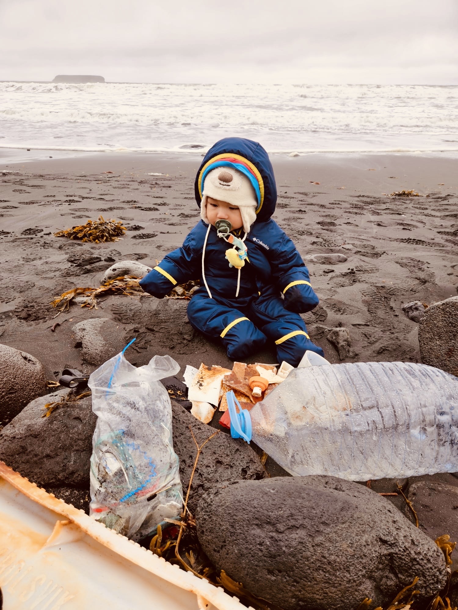 A toddler sitting on a beach in Iceland in winter overalls and a beanie that has bear ears. The child looks at plastic bottles on the sand.