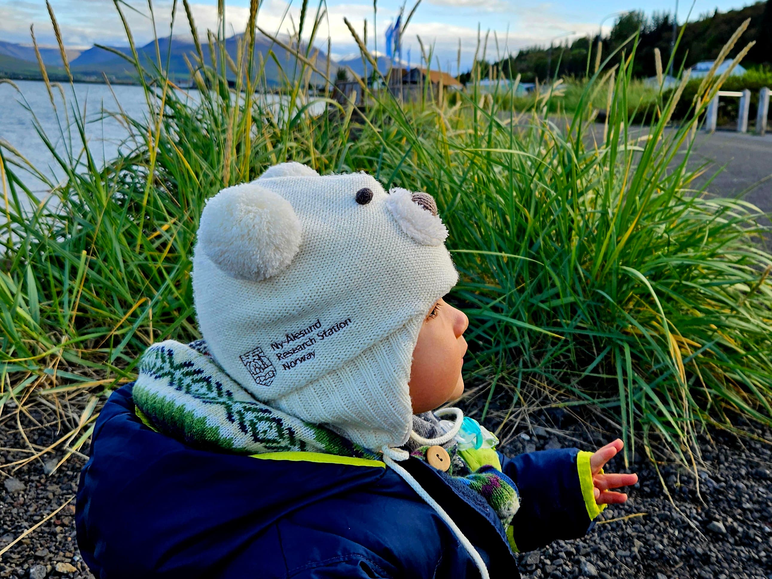 A toddler sitting on an Arctic beach with winter overalls and a beanie that has bear ears and text "Ny-Ålesund Research Station Norway".