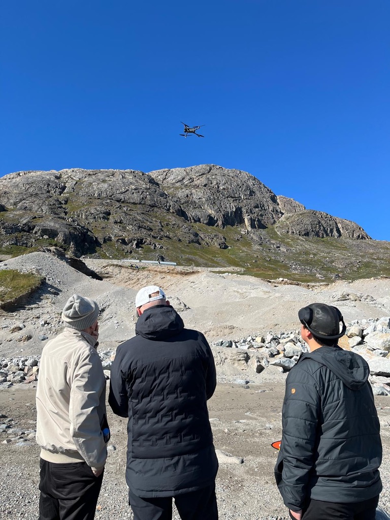 Three people flying a drone on a rocky beach with a remote control.