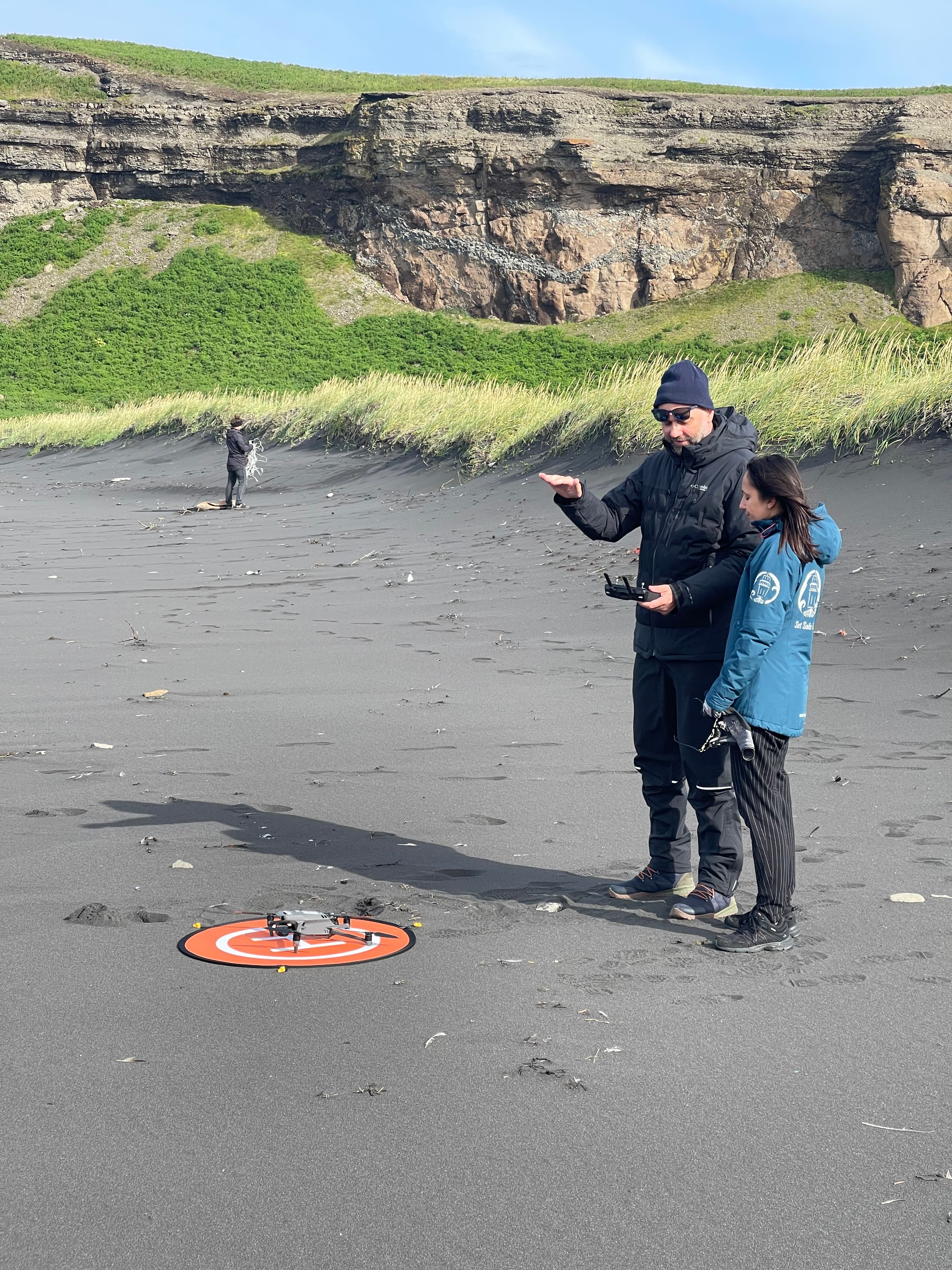An older person and a youngster standing next to a drone laying on the ground on an orange platform.