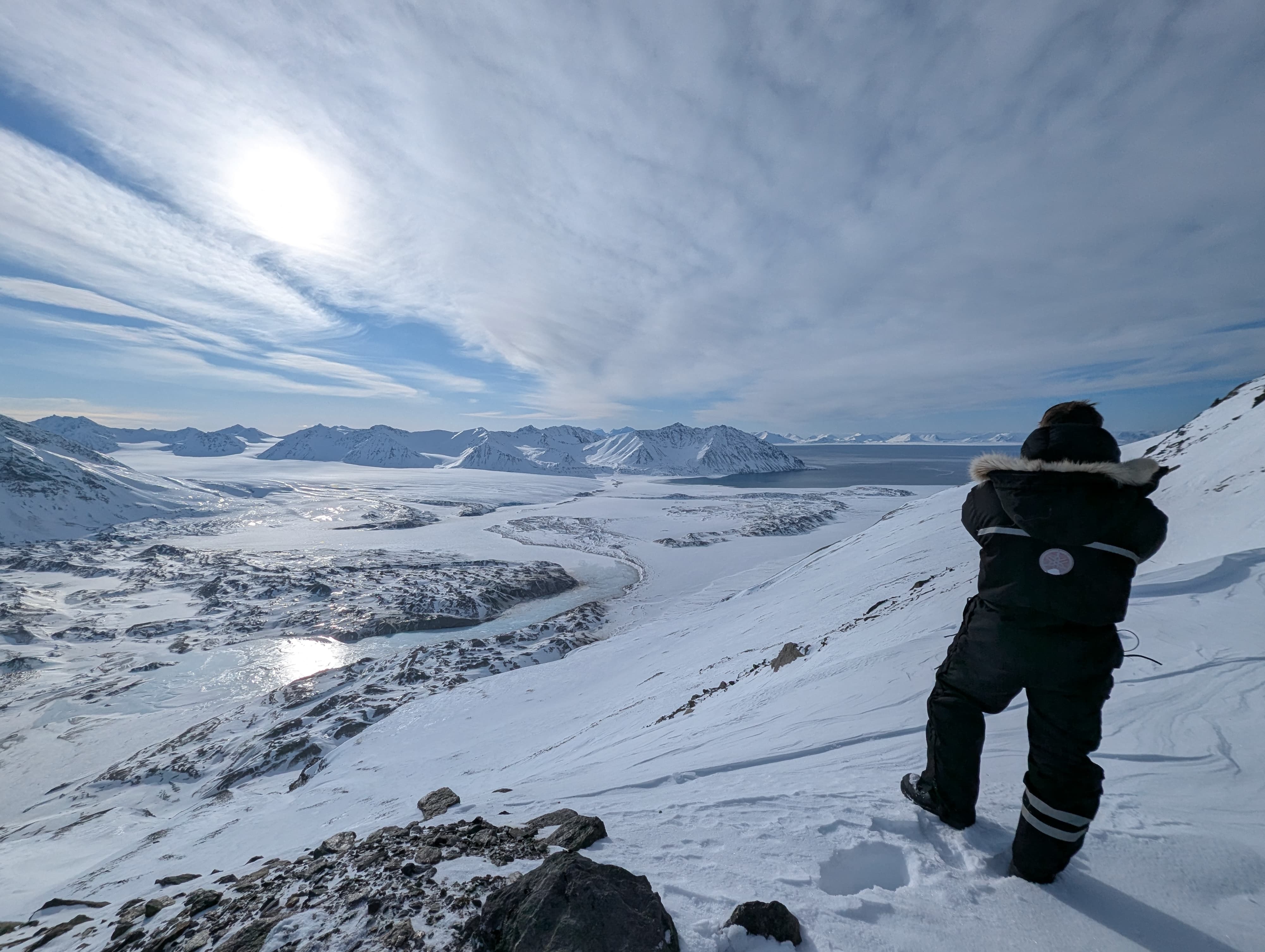 A researcher in full winter clothing stands before a wide snowy glacier. In the horizon are snowy mountains.