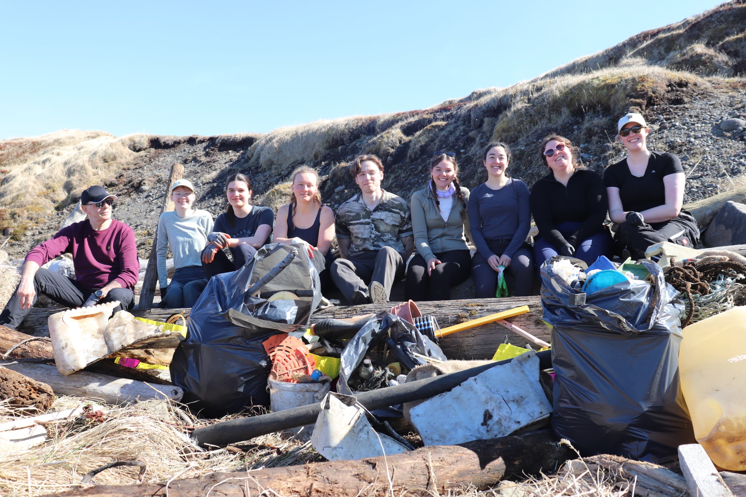 Volunteers sitting on a row, smiling for the camera. In front of them is a large pile of plastic litter they have collected from the beach.