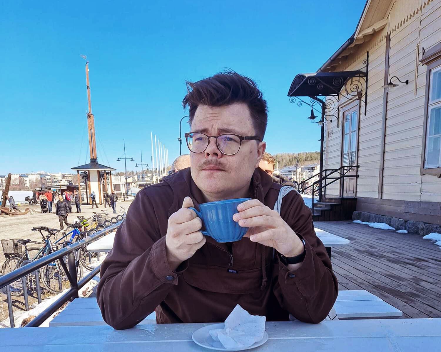 Ilkka Olkku sits by an outside coffee table holding a cup and looks away from the camera.