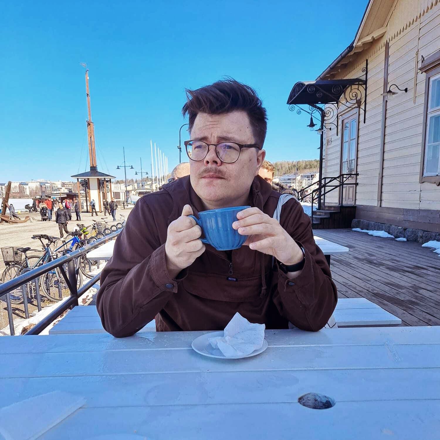 Ilkka Olkku sits by an outside coffee table holding a cup and looks away from the camera.