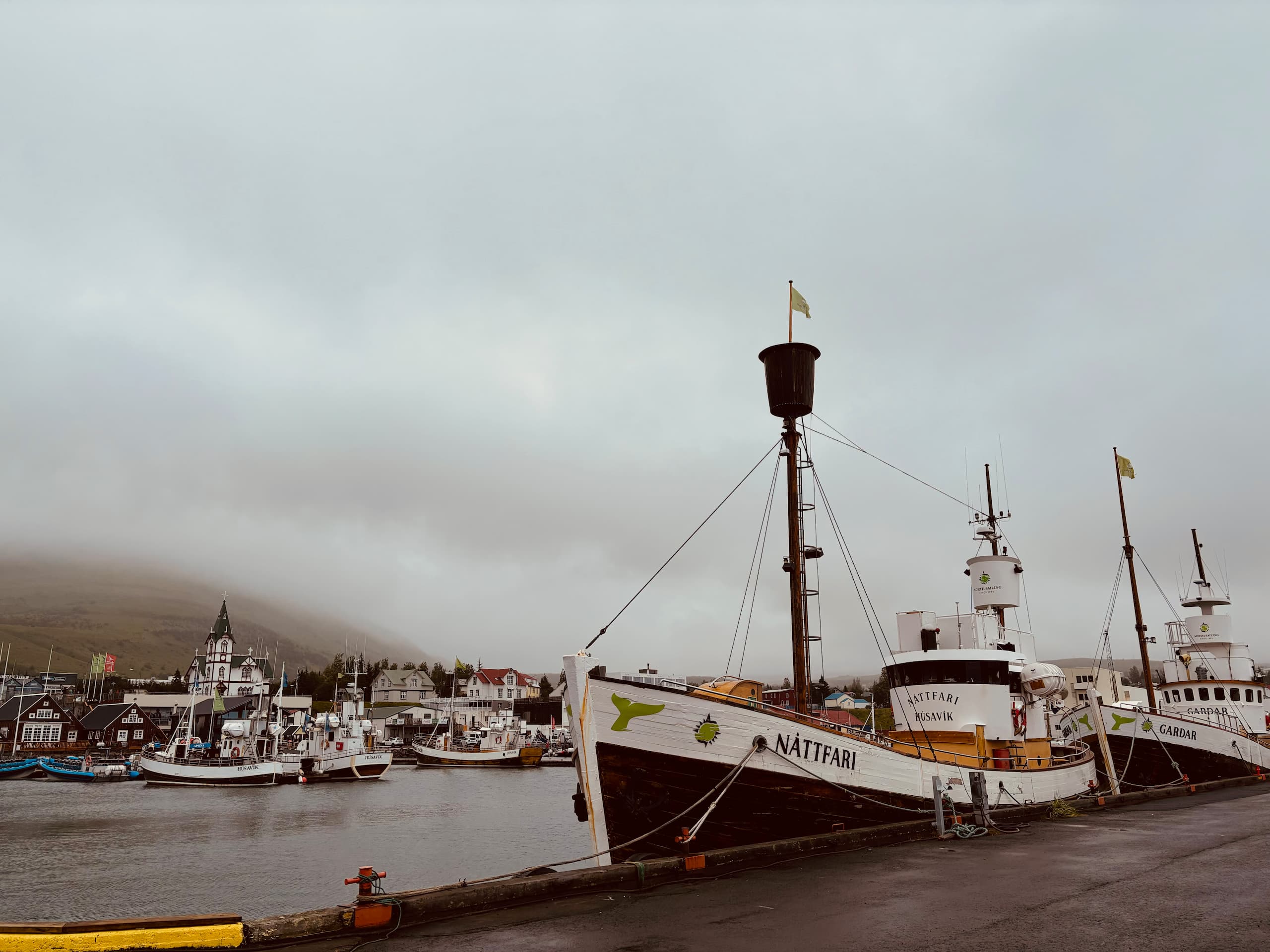 A ship named Náttfari in the harbor of Húsavík. Small houses and a misty mountain in the background.
