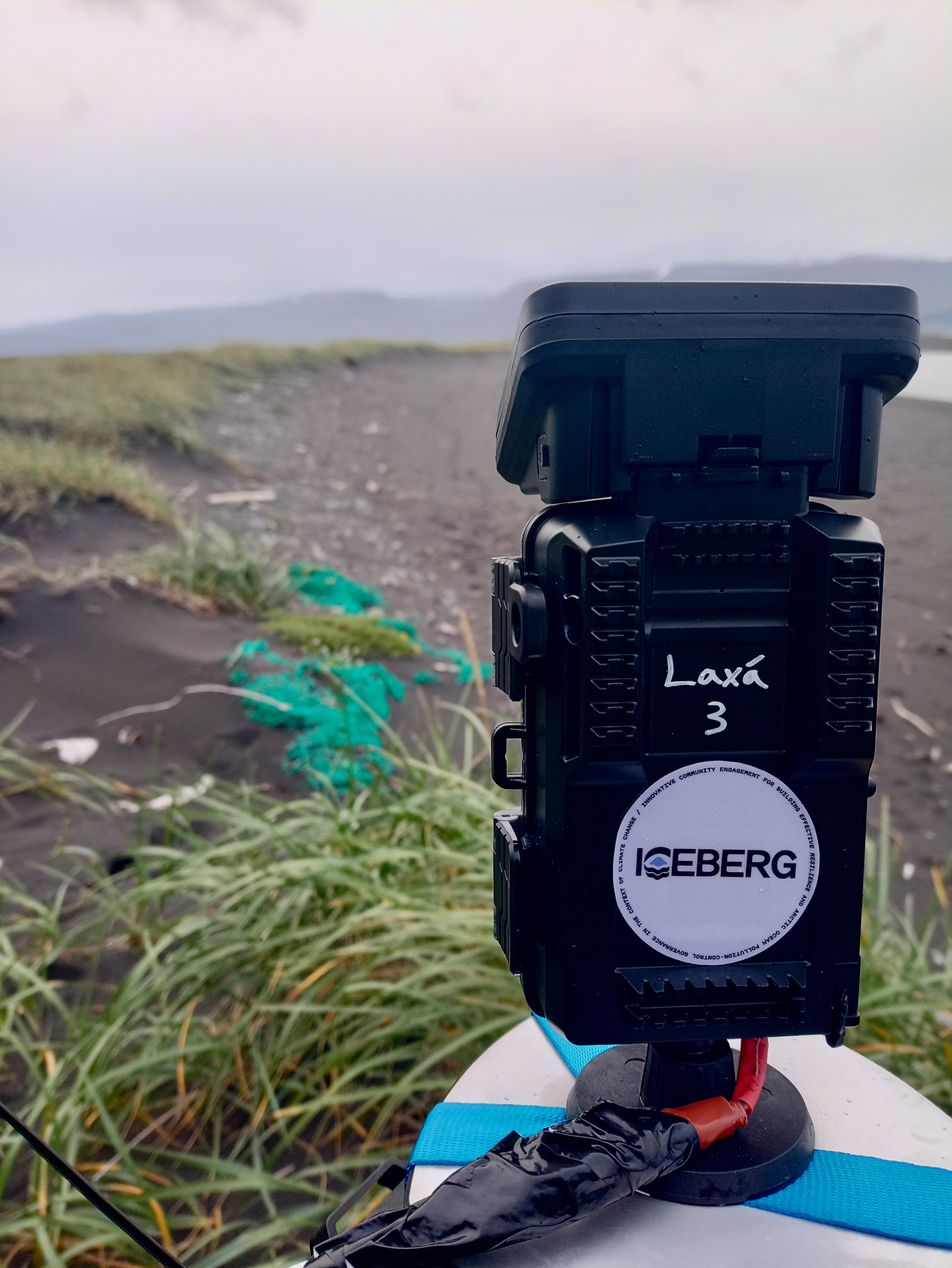 A close-up of a time-lapse camera on a rocky beach. The camera has an ICEBERG sticker on it.