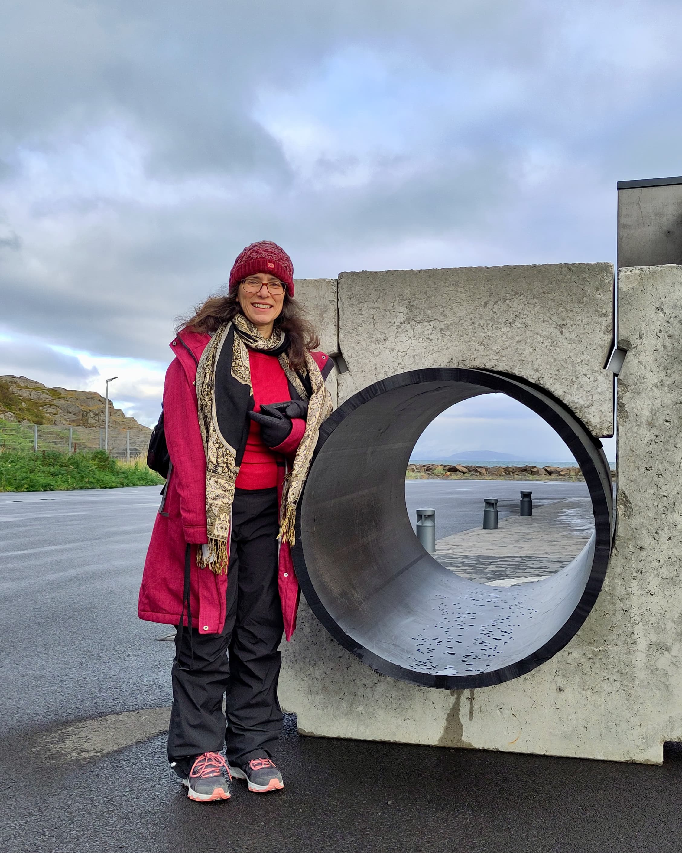 Christa Marandino stands next to a concrete element that has a hole in it. She is in outdoor clothing and there's a fjord in the background.