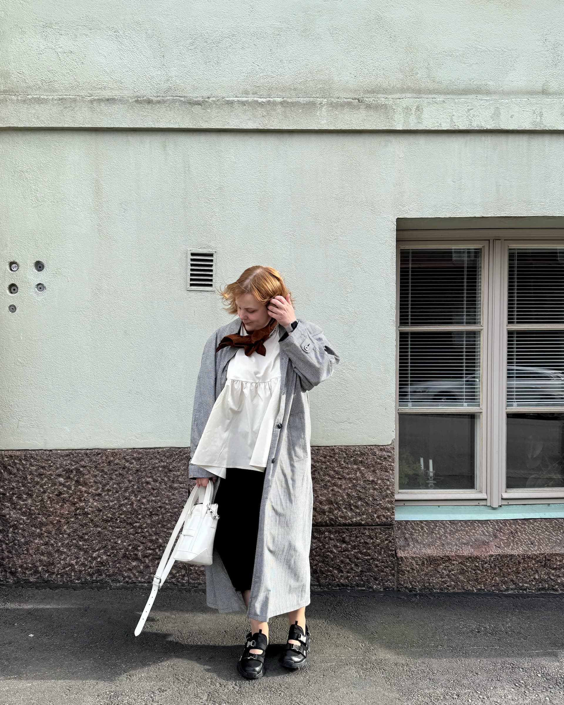 Janika Lähdes stands in front of a wall, looks down and moves her hair behind her ear.