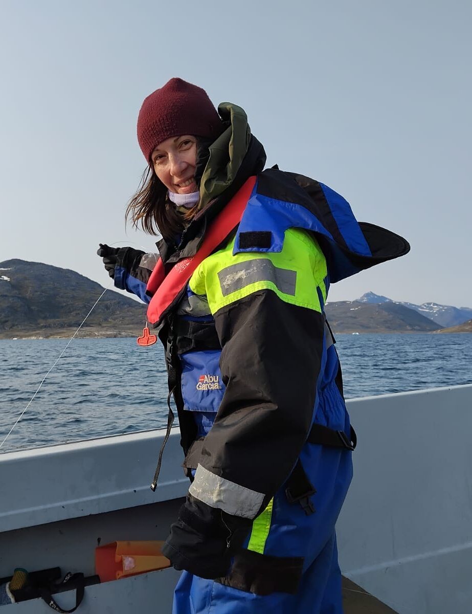 Project manager Élise Lépy wearing cold-weather gear and a life jacket stands on a small boat at sea, holding a line over the water, with rocky Arctic coastline and mountains in the background.