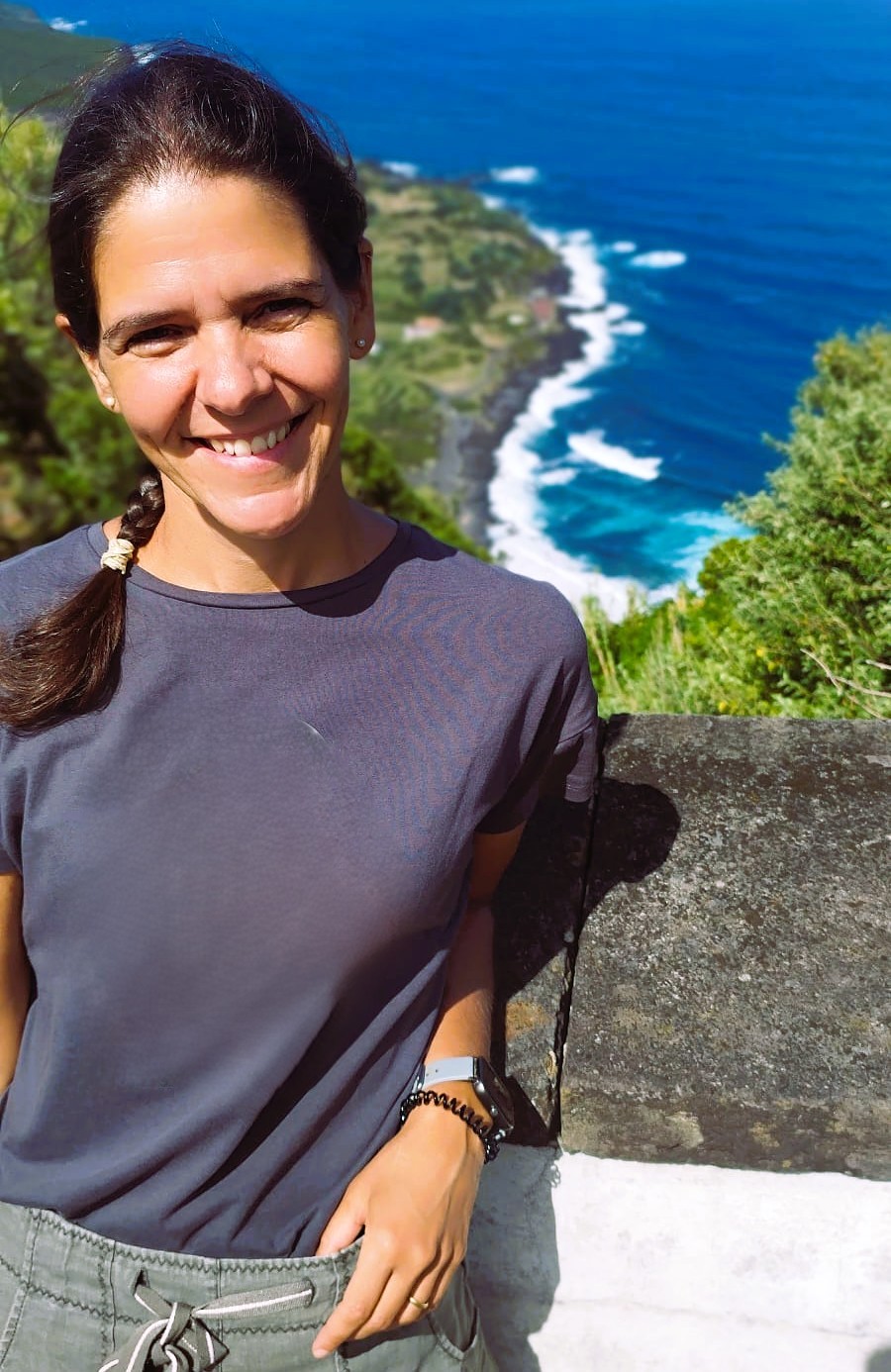 Ana Faria standing outdoors by a coastal landscape, smiling, with the ocean and rugged shoreline in the background.