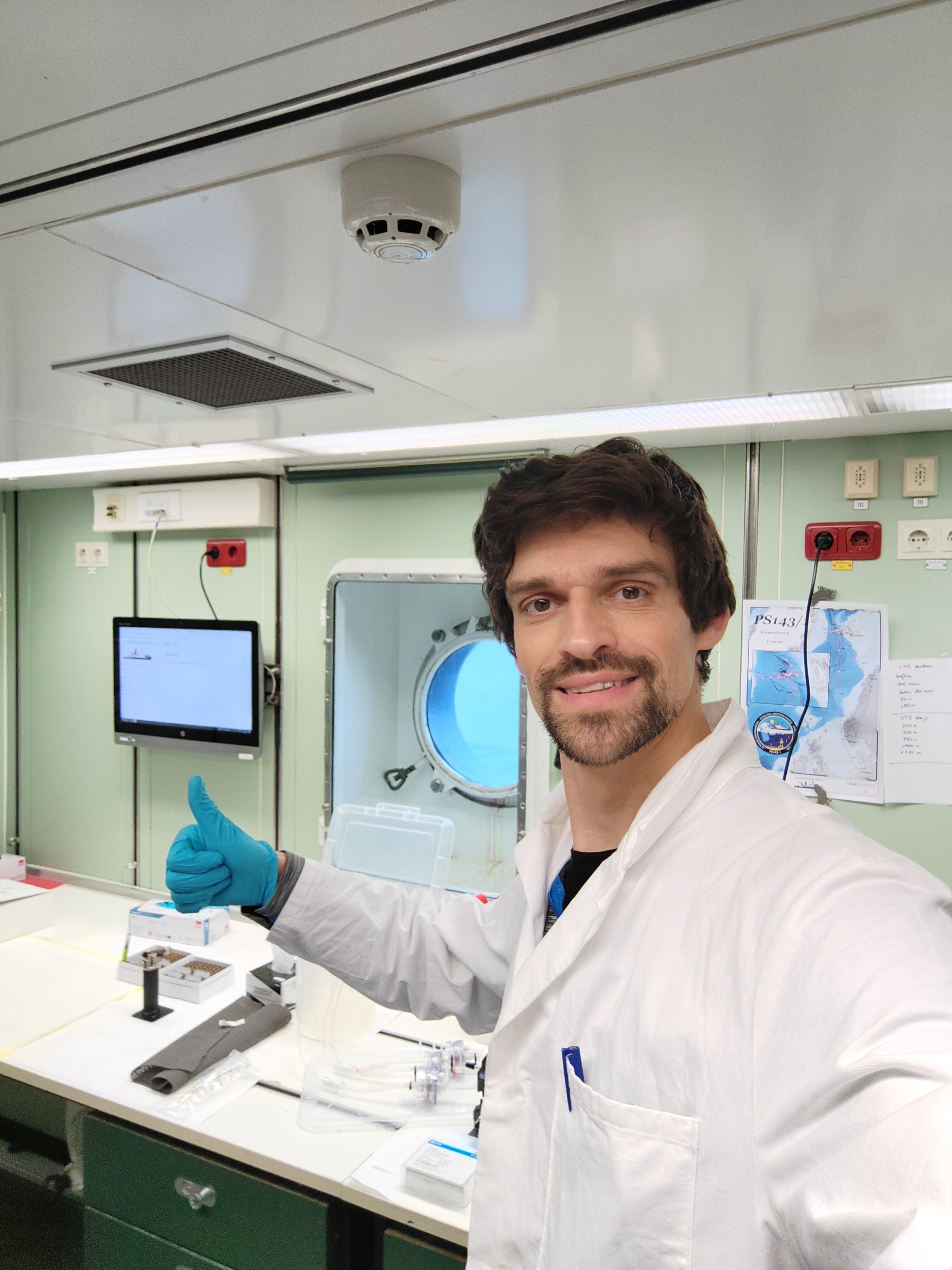 Picture of a dark featured man smiling. He is wearing a lab coat and gloves, in a lab setting.