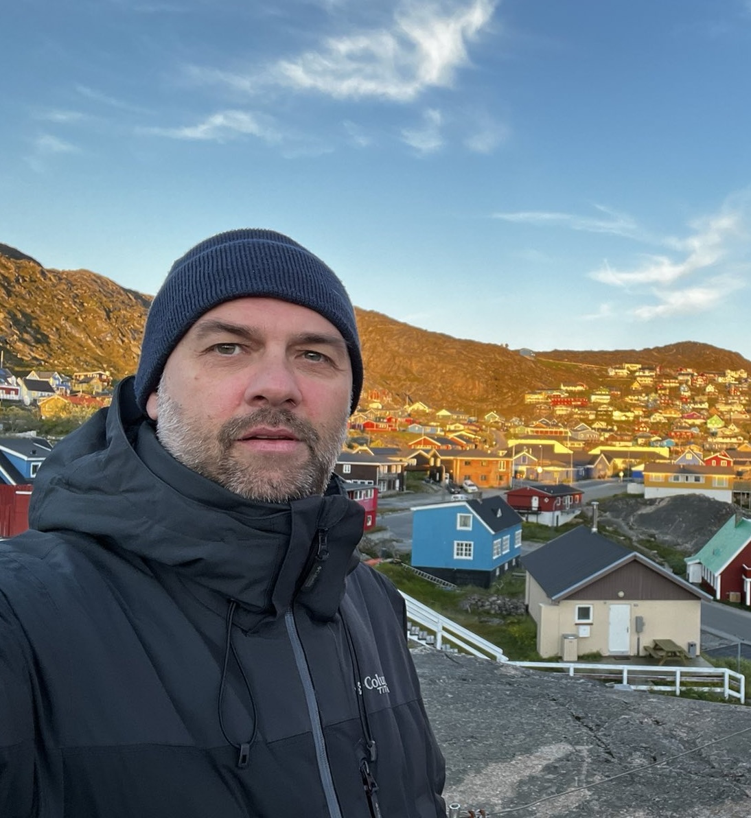 A bearded man standing in front of a landscape of colorful houses, lit up by the sun. A blue sky can also be seen.
