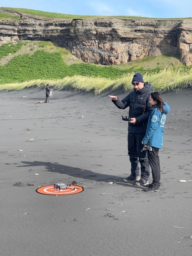 Two people dressed in outerwear standing next to a drone on the ground, presumably about to fly it. The ground is a dark grey color.