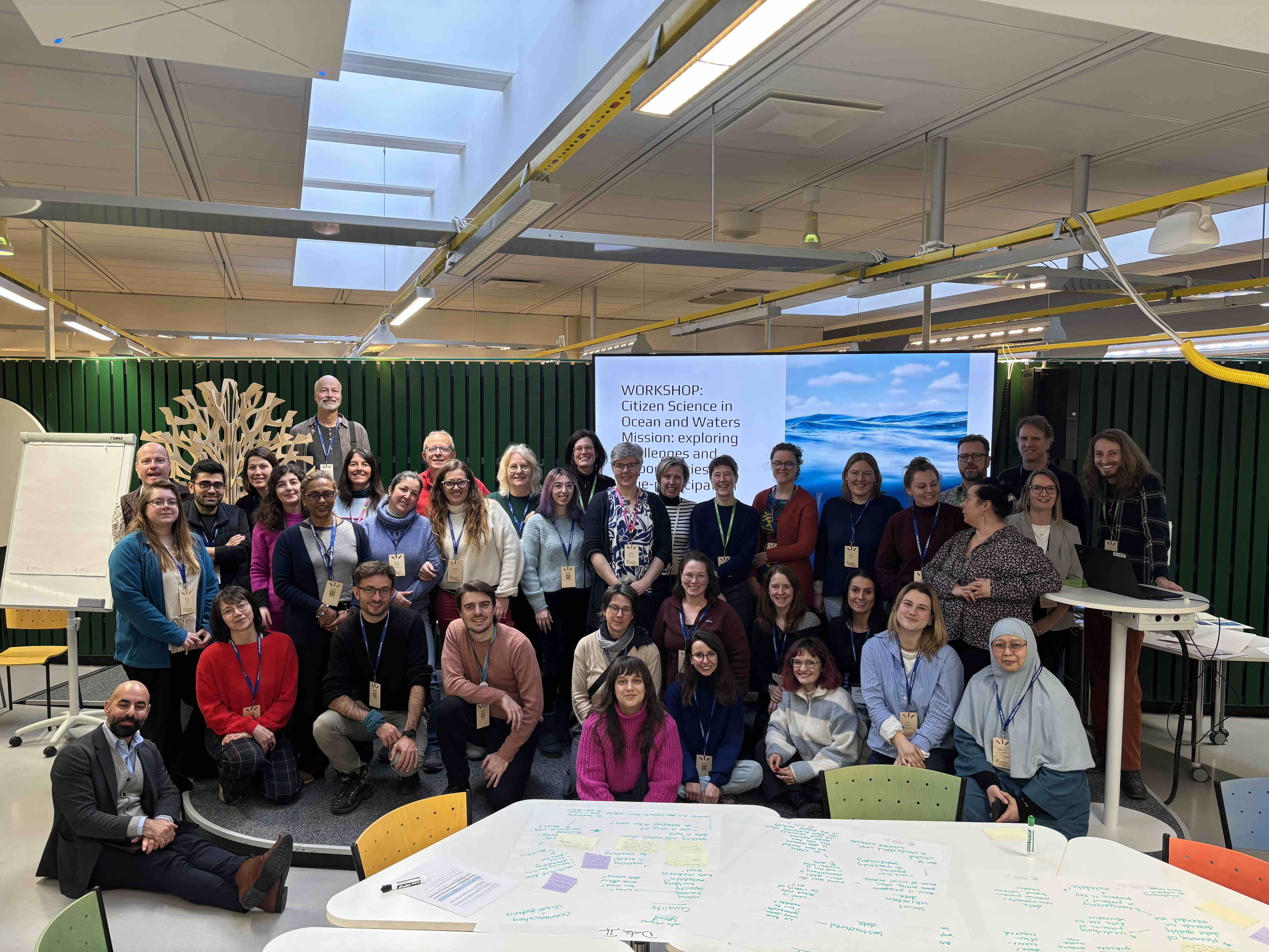 A group of smiling people standing together in a colorful setting at the University of Oulu campus. Behind them is a screen showcasing the text "Citizen science in Ocean and Waters Mission".