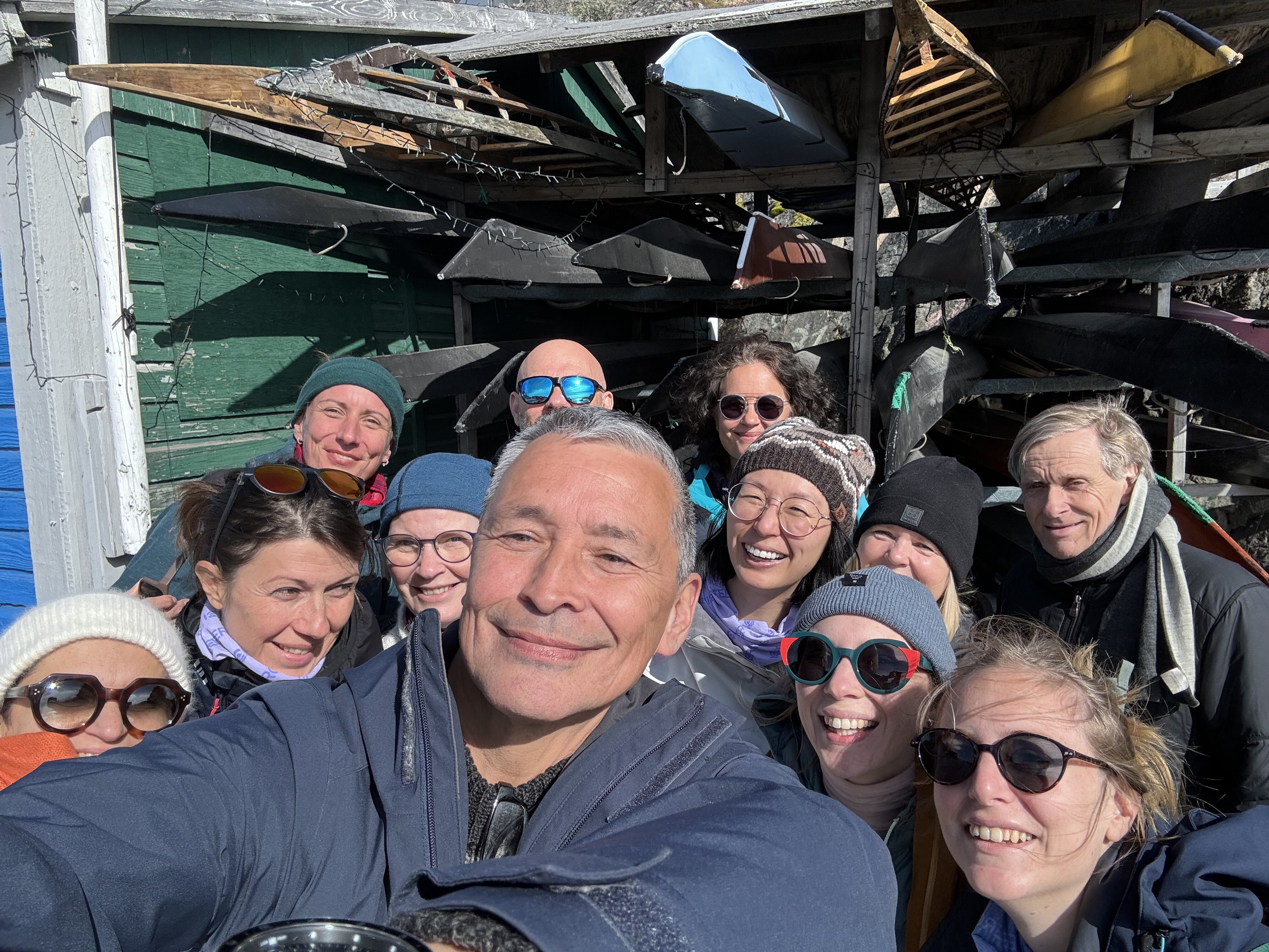A group selfie with about 12 people smiling and looking happy. They are standing outside in the sun, some are wearing sunglasses and hats.