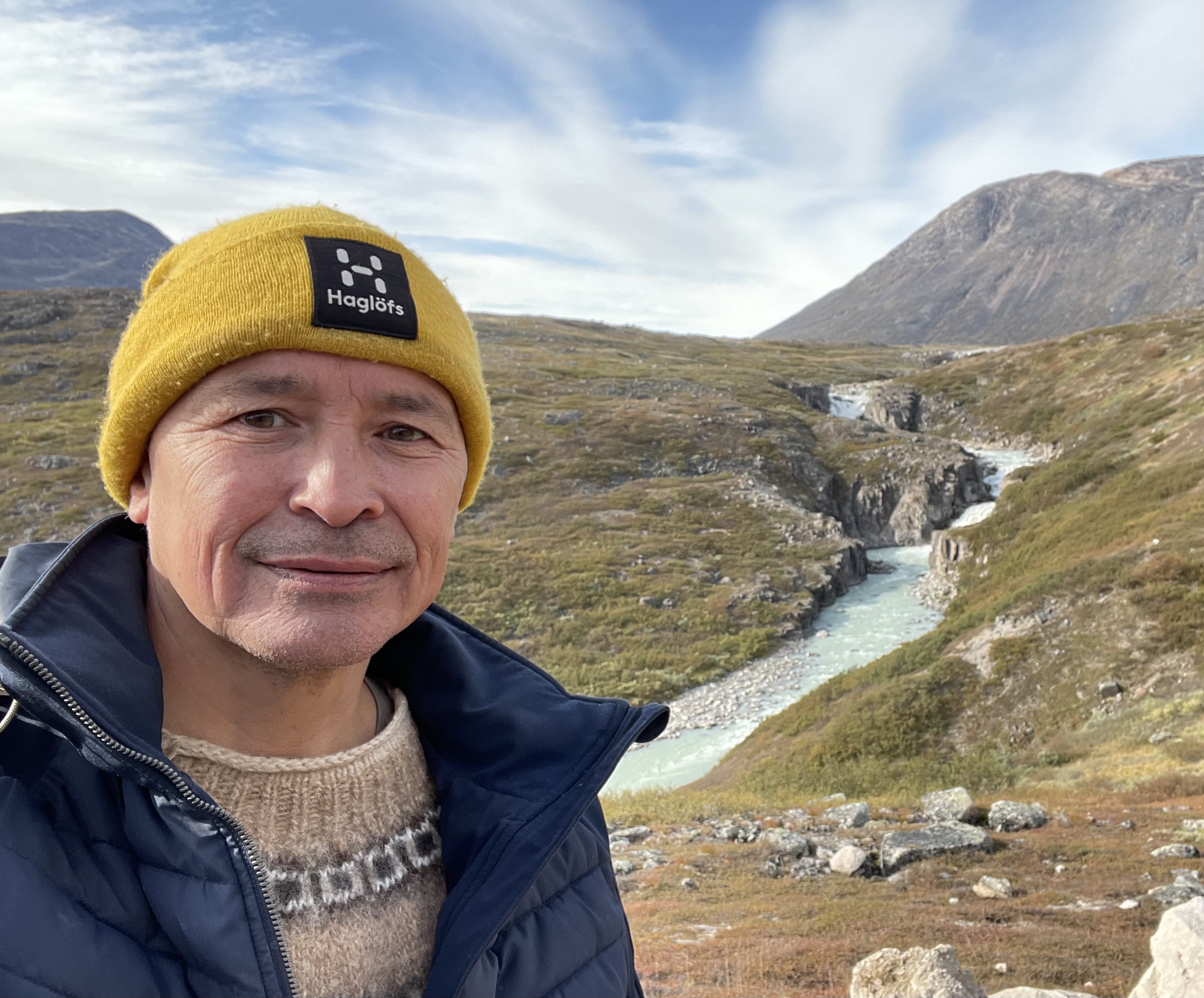 A man with a yellow hat standing in front of a mountain scenery with a waterfall. A blue sky can be seen in the distance.