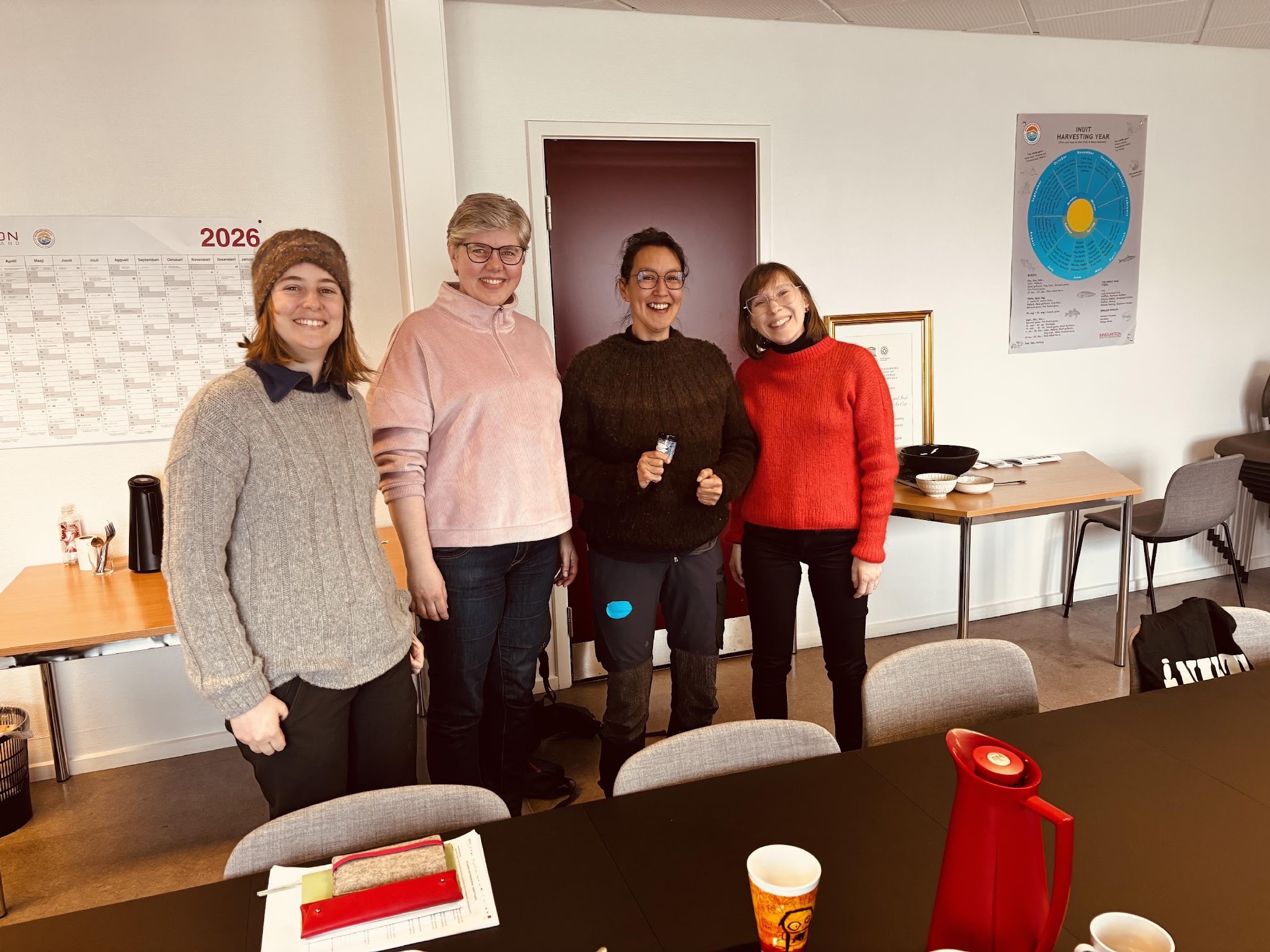 Four women standing in a row and smiling. Next to them is a big table with coffee and other things on it.