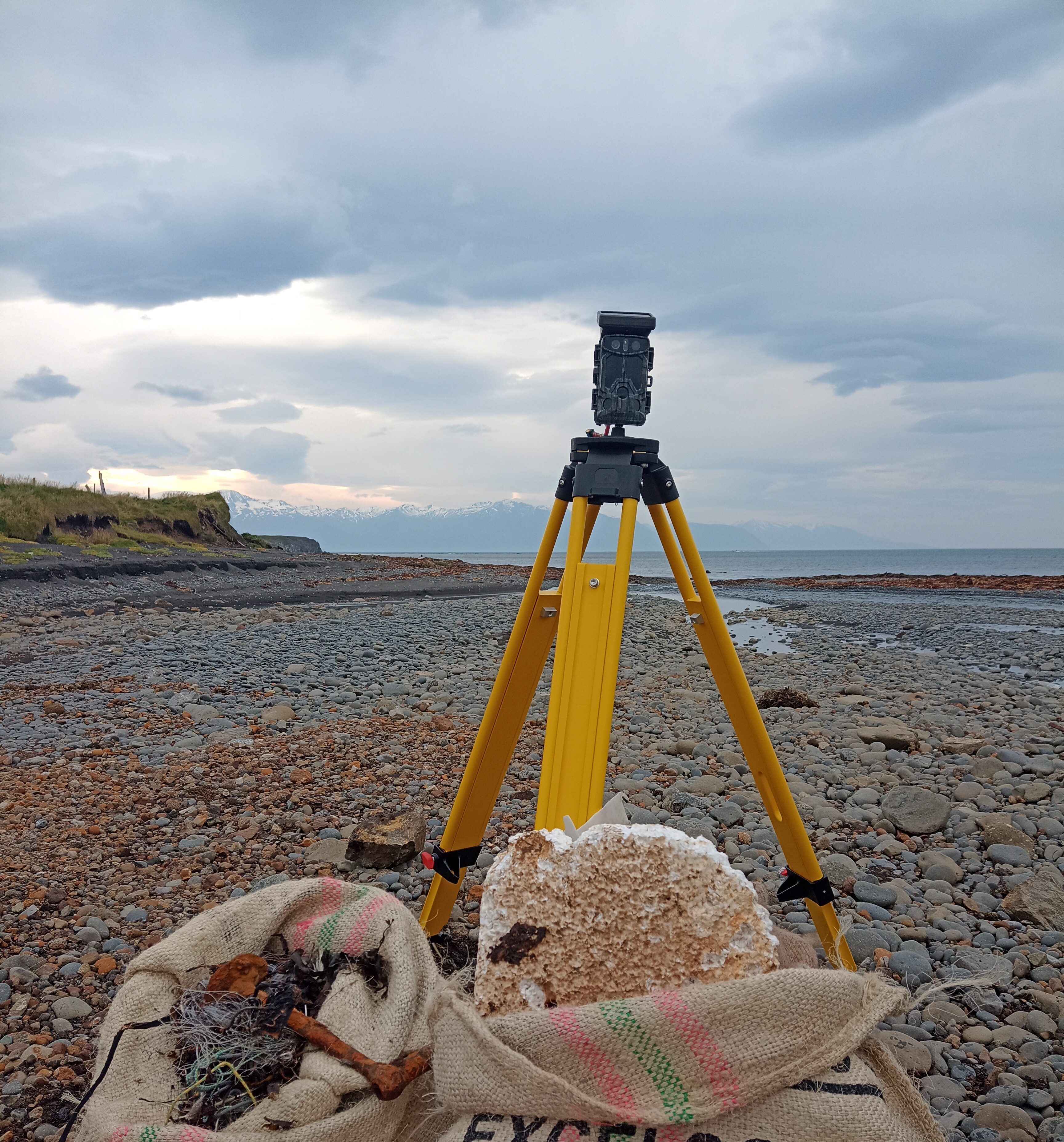 A small, black camera on a bright yellow stand, on a rocky beach. In the background is a gloomy and cloudy landscape.