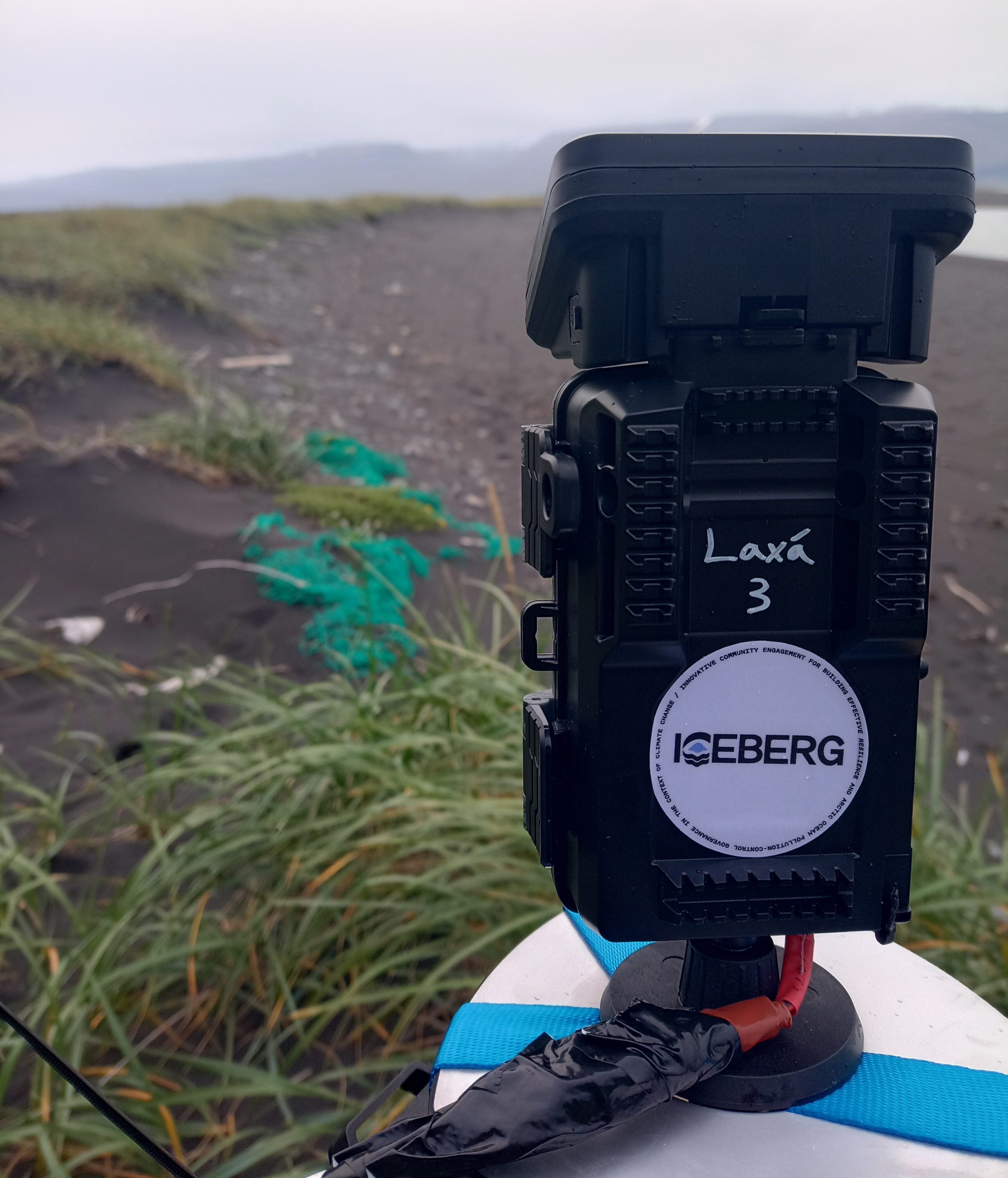 Close up image of a black time-lapse camera with a light purple sticker with the ICEBERG project logo on it. In the background, a gloomy beach landscape.