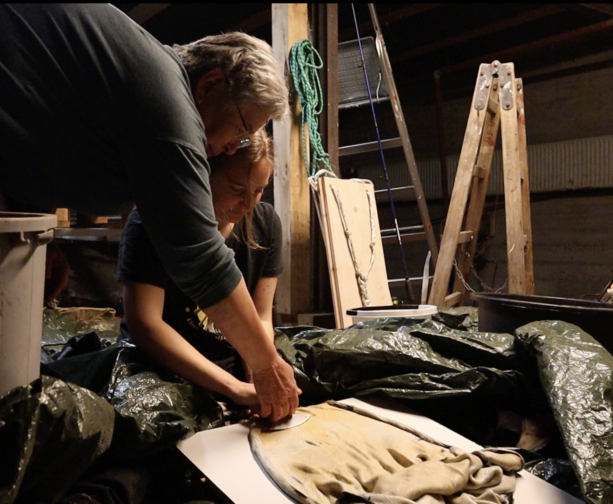 Two people hunched over a piece of seal skin, using some type of tool on it. They are sitting in a dark, shed-type building.