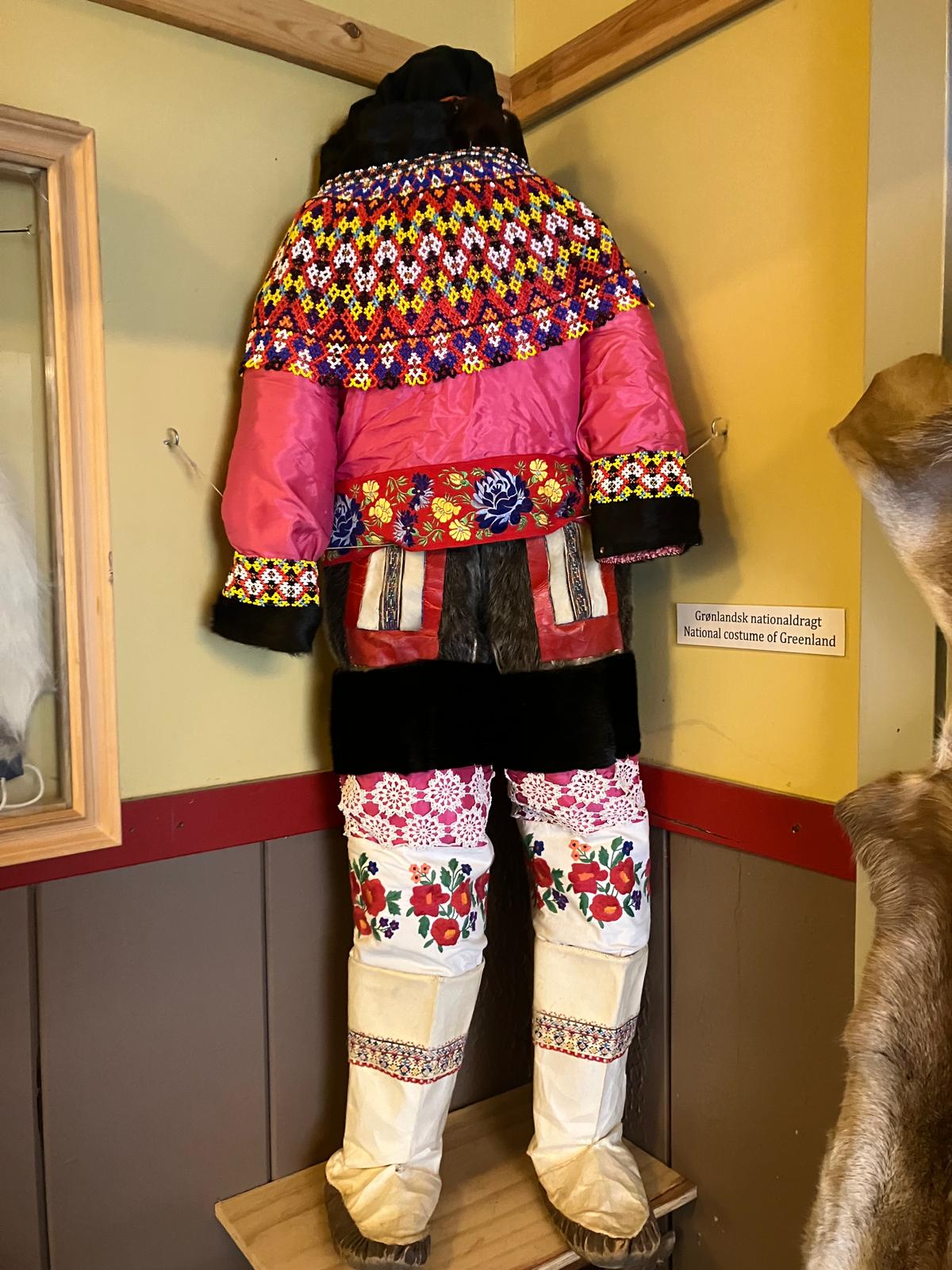 A colorful national costume hanging from the ceiling against a yellow wall. It's mostly pink, with variations of yellow, white, green and other colors.
