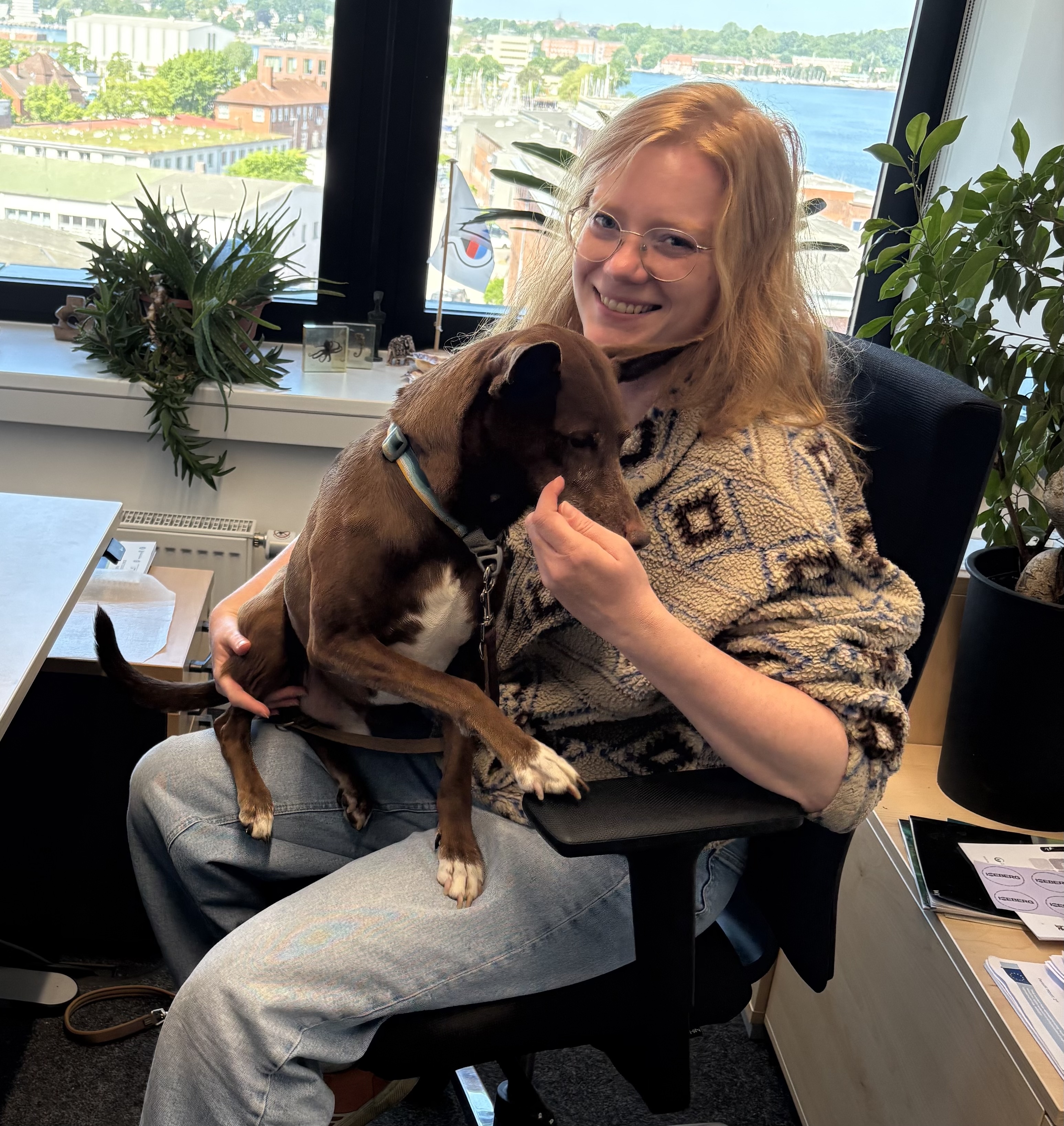 A woman with light long hair and glasses, sitting in an office chair and smiling. She has a small brown dog in her lap.
