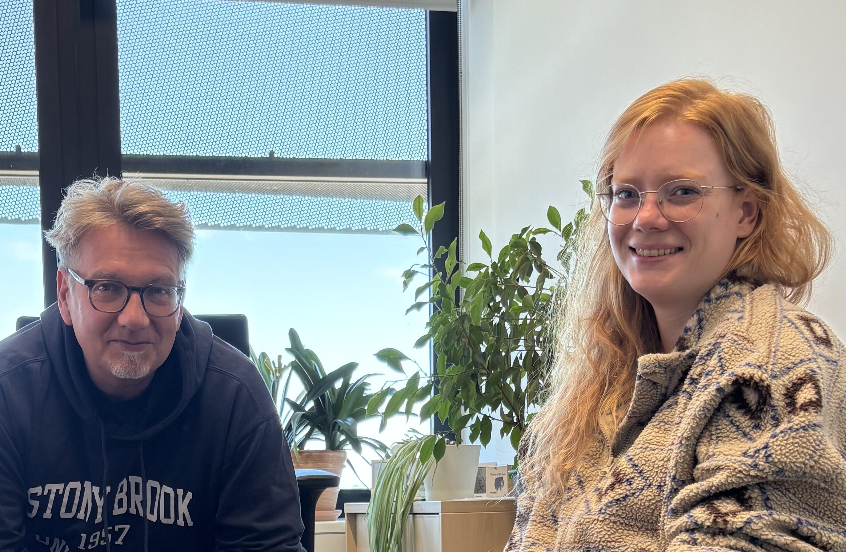 Two people sitting and smiling in an office setting. In the right down corner there is a brown dog with pointy ears.