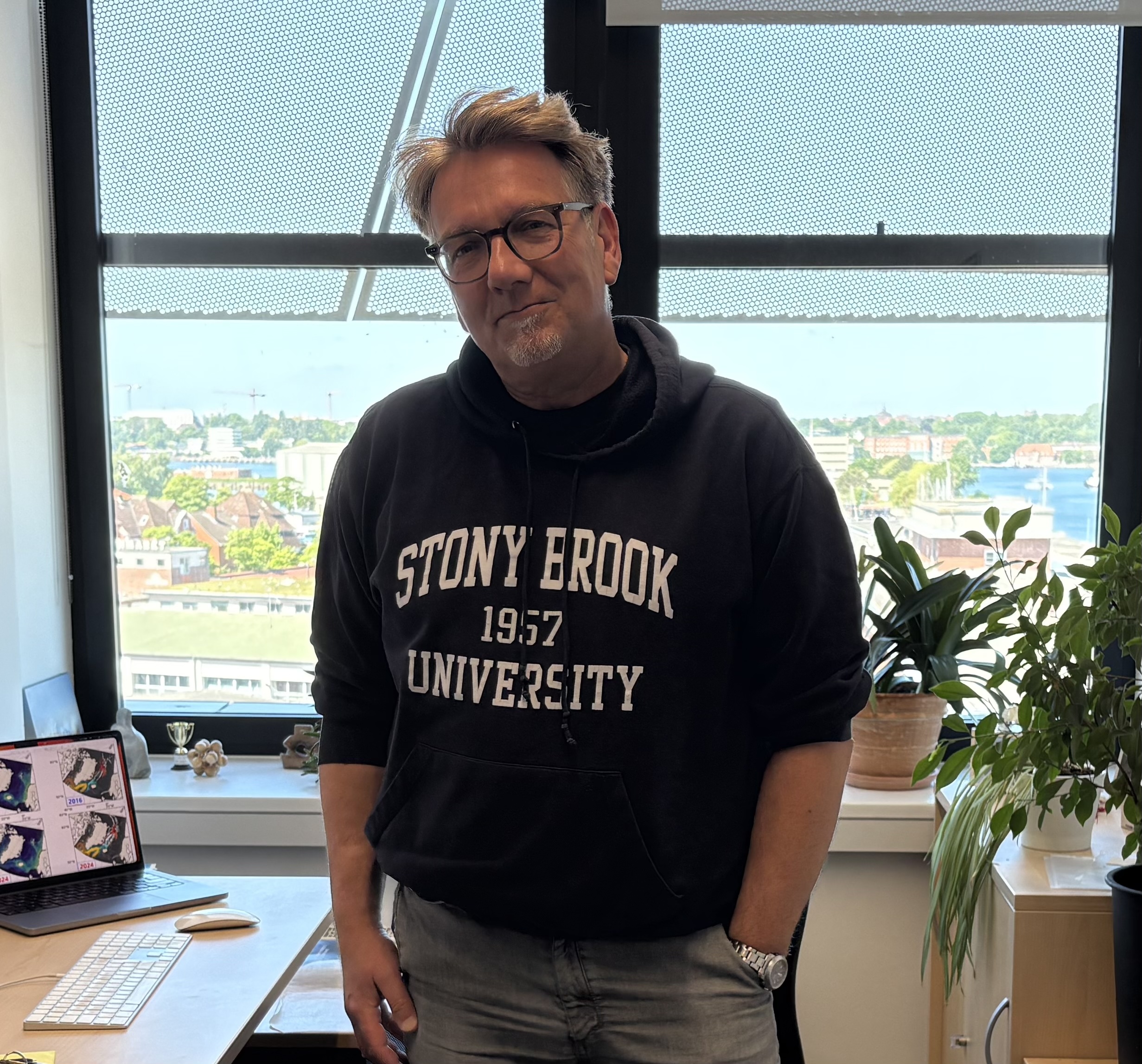 A man with light brown hair and glasses standing and smiling in an office setting. He is wearing a dark hoodie with the text "Stony brook 1957 university" on it.
