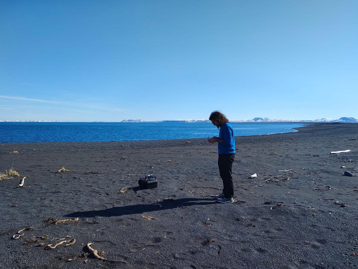 A person flying a drone on a beach. The sand is dark brown and the blue shoreline can be seen in the background, as well as the blue sky. The person flying the drone has a blue shirt.