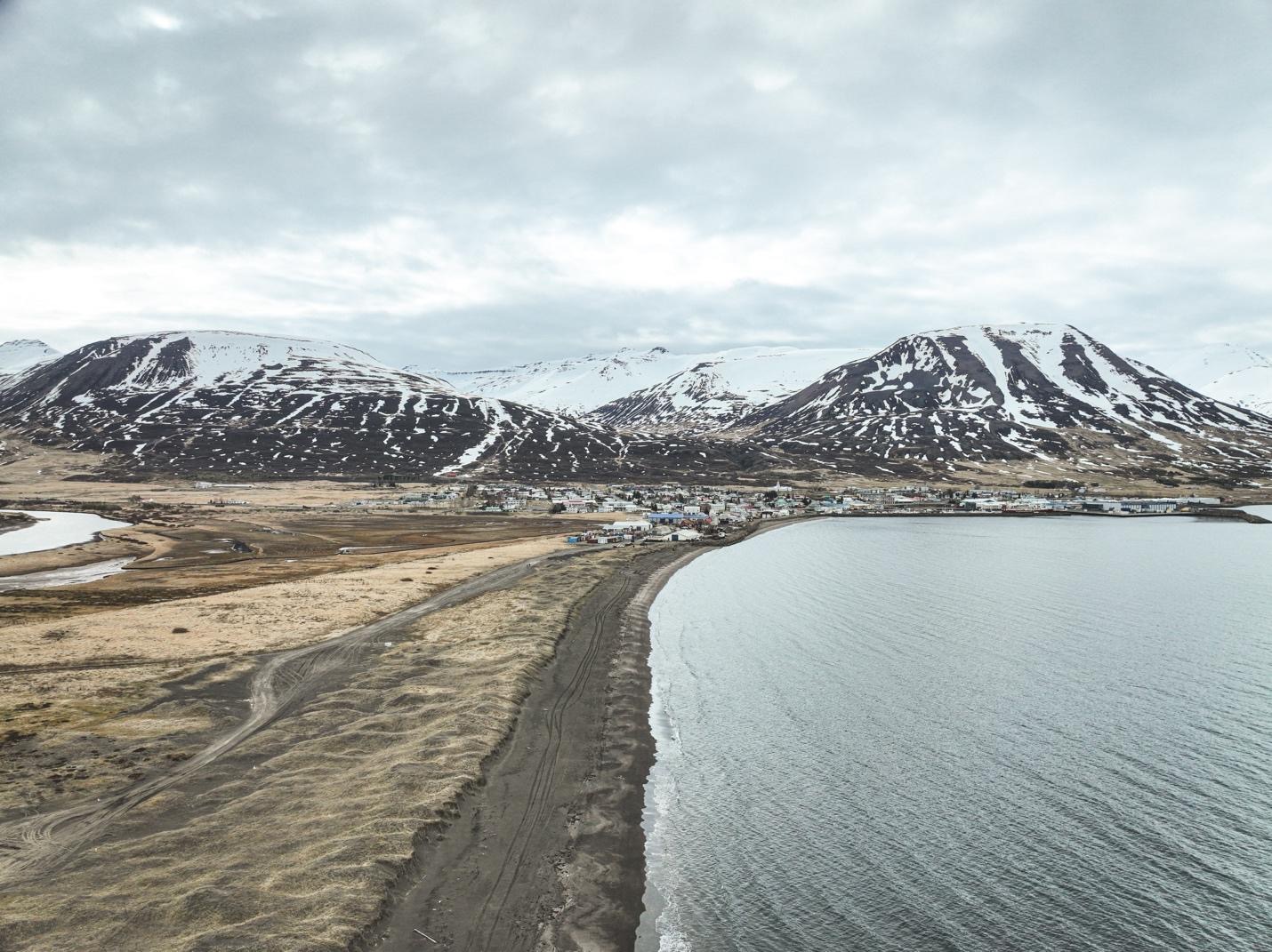 Two large snow-covered mountains and a small village in front of them. The village sits on the shore of a large body of water