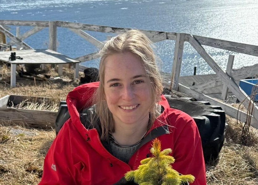 A smiling woman with light hair in a red hoodie, sitting in a squat and holding a small tree seedling.