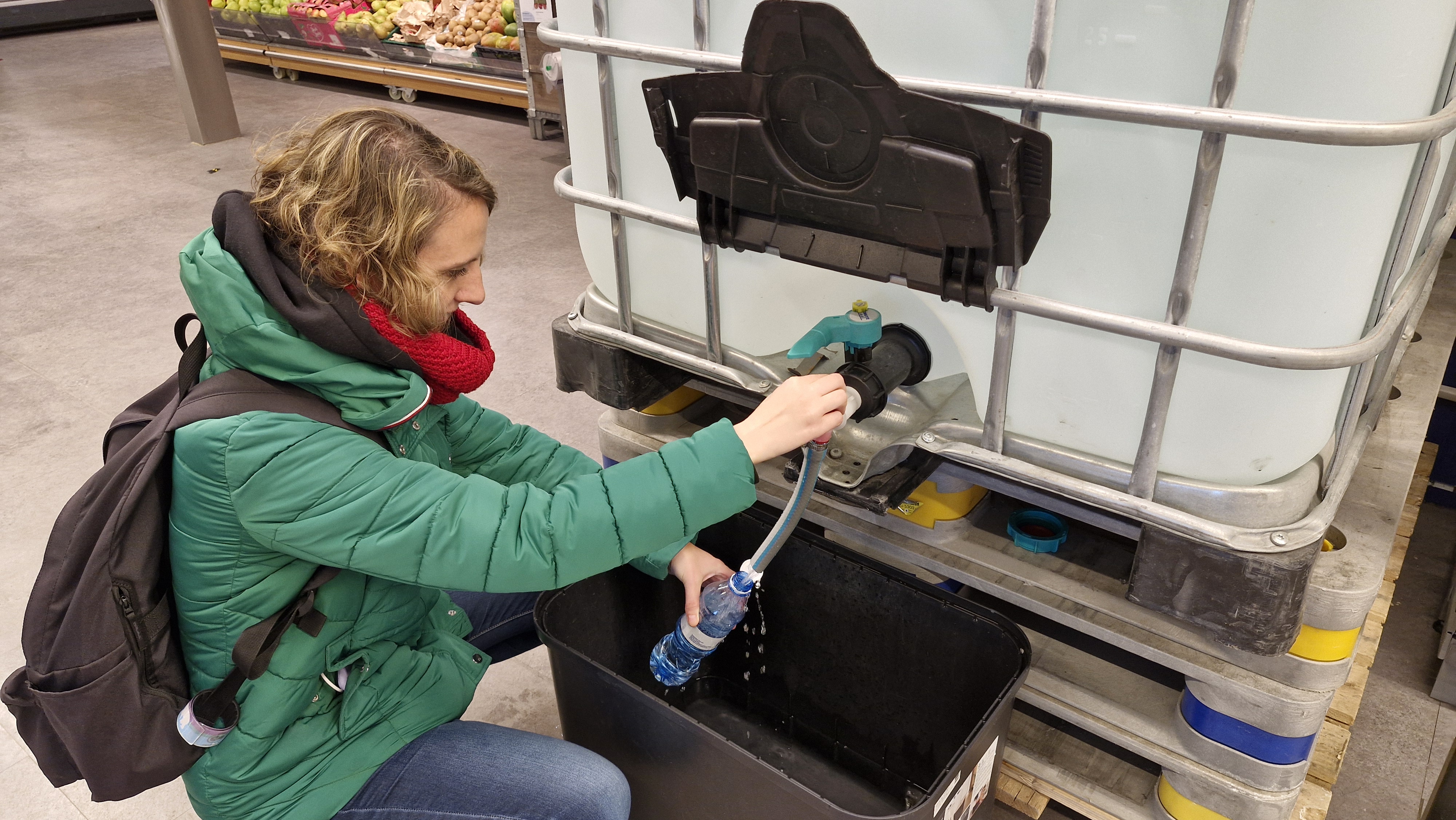 A woman in a green jacket pouring water into her water bottle from a large industrial water container.