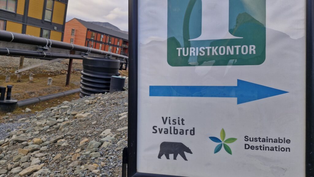 A sign that says "Visit Svalbard" with the picture of a bear, and "Sustainable destination" with a blue arrow. Behind sign, there are water pipes.