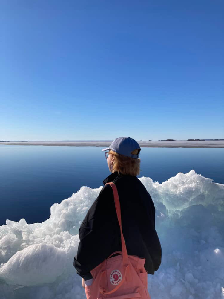 A woman with short hair, a blue hat, and a pink backpack standing with her back against the camera. She is standing in a snowy landscape, looking out towards the sea.