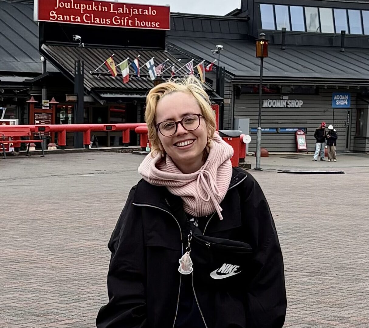 A woman with light, short hair and dark glasses smiling at the camera. In the background, there is a building with the text "Santa claus gift shop".