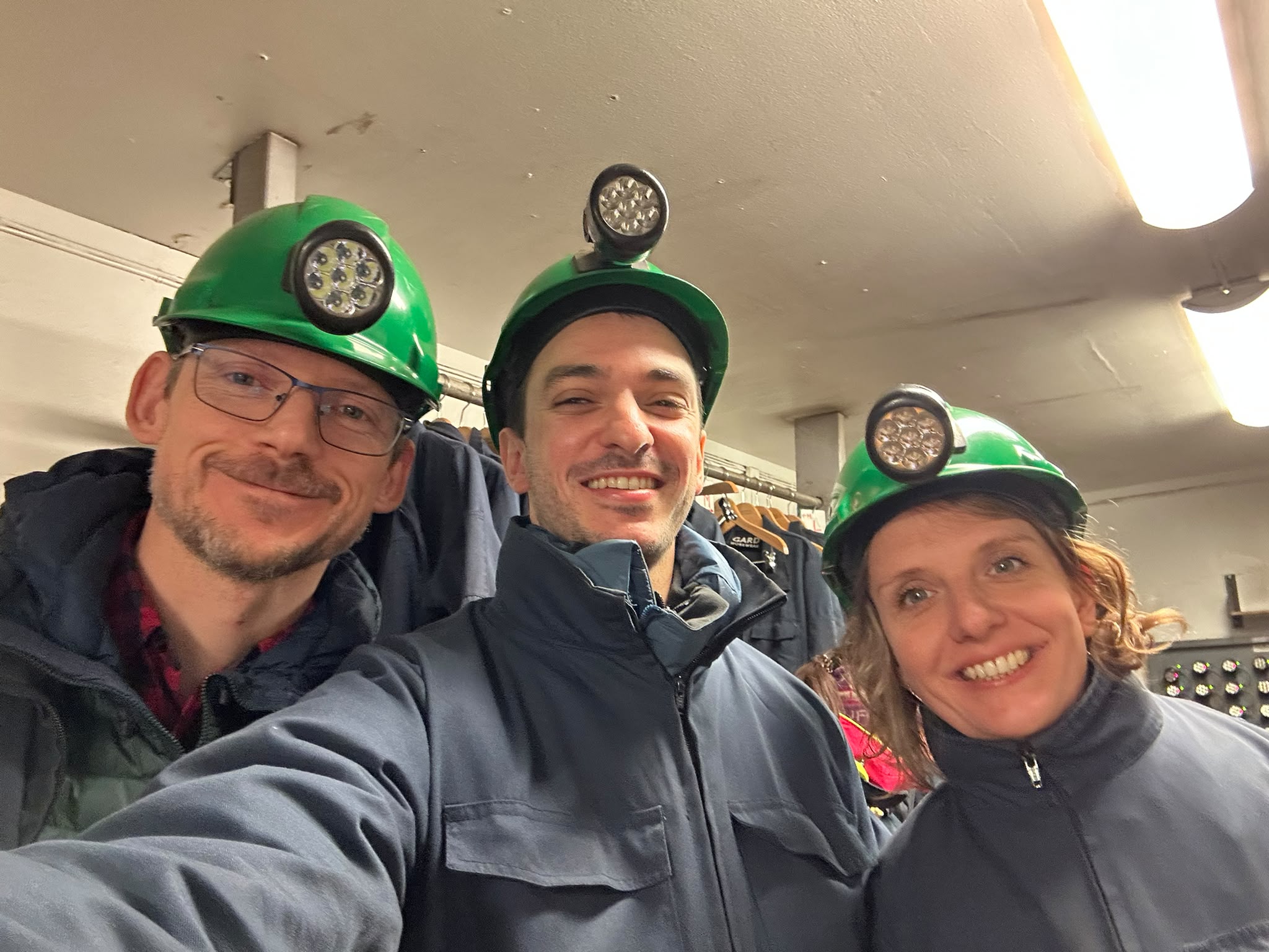 Selfie of three people smiling, wearing green hard hats with lamps on them and dark jackets.