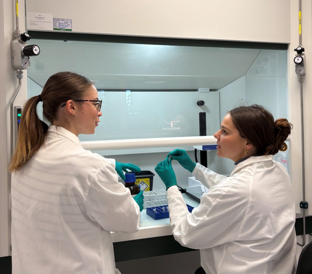 Two women in lab coats talking and performing tests in a laboratory setting. One of them is standing and the other is sitting. They are in the middle of a conversation.