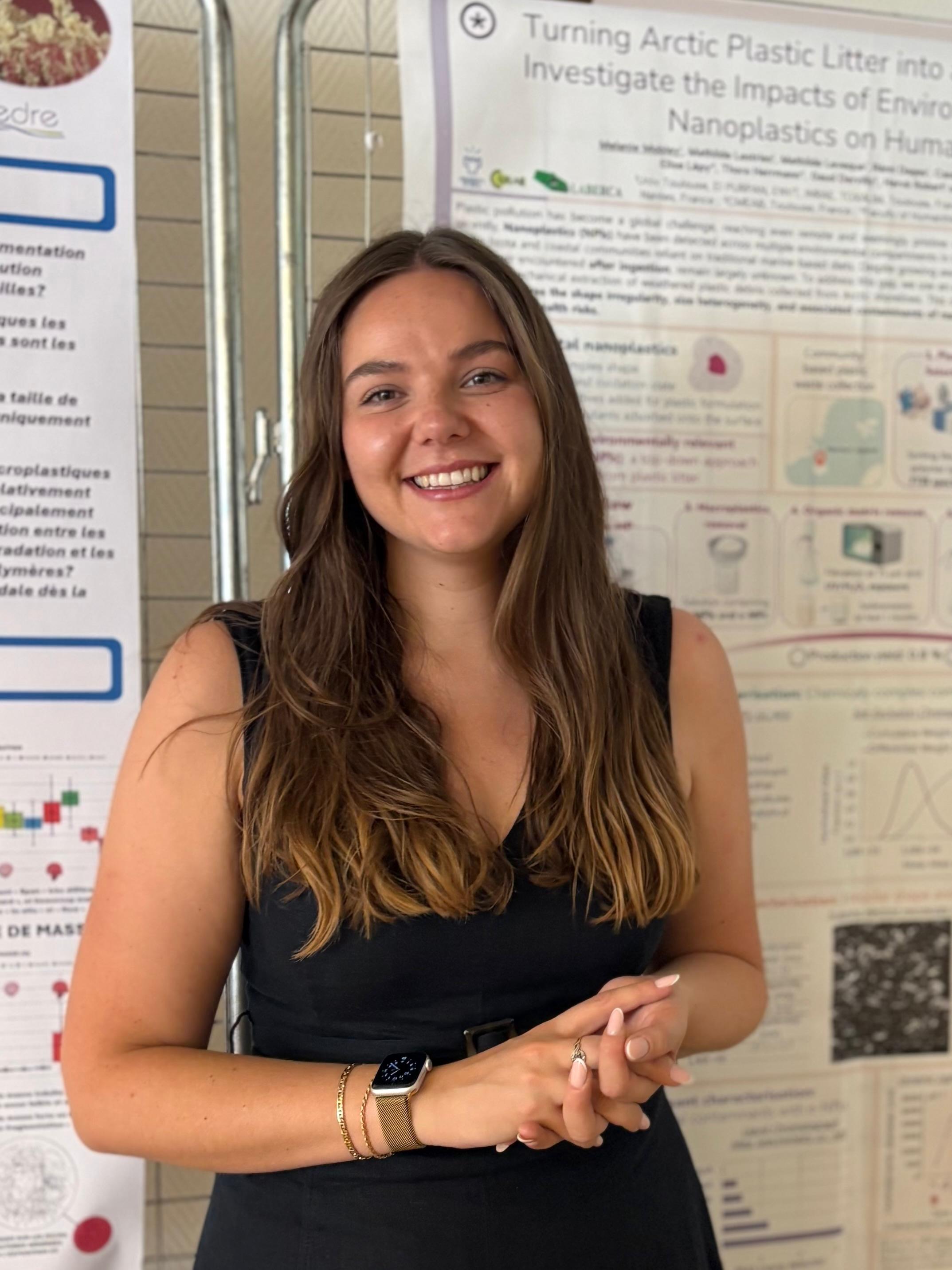 A smiling woman with long, brown hair and a black shirt standing in front of research posters.