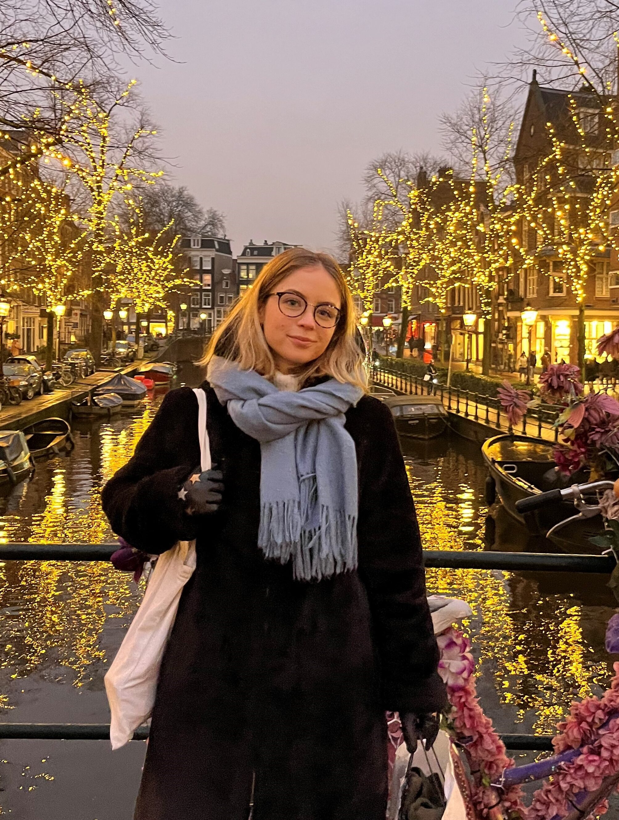 A woman with glasses and light, short hair wearing a black jacket and light blue scarf. She is standing on a bridge, with water and houses with fairy lights behind her.